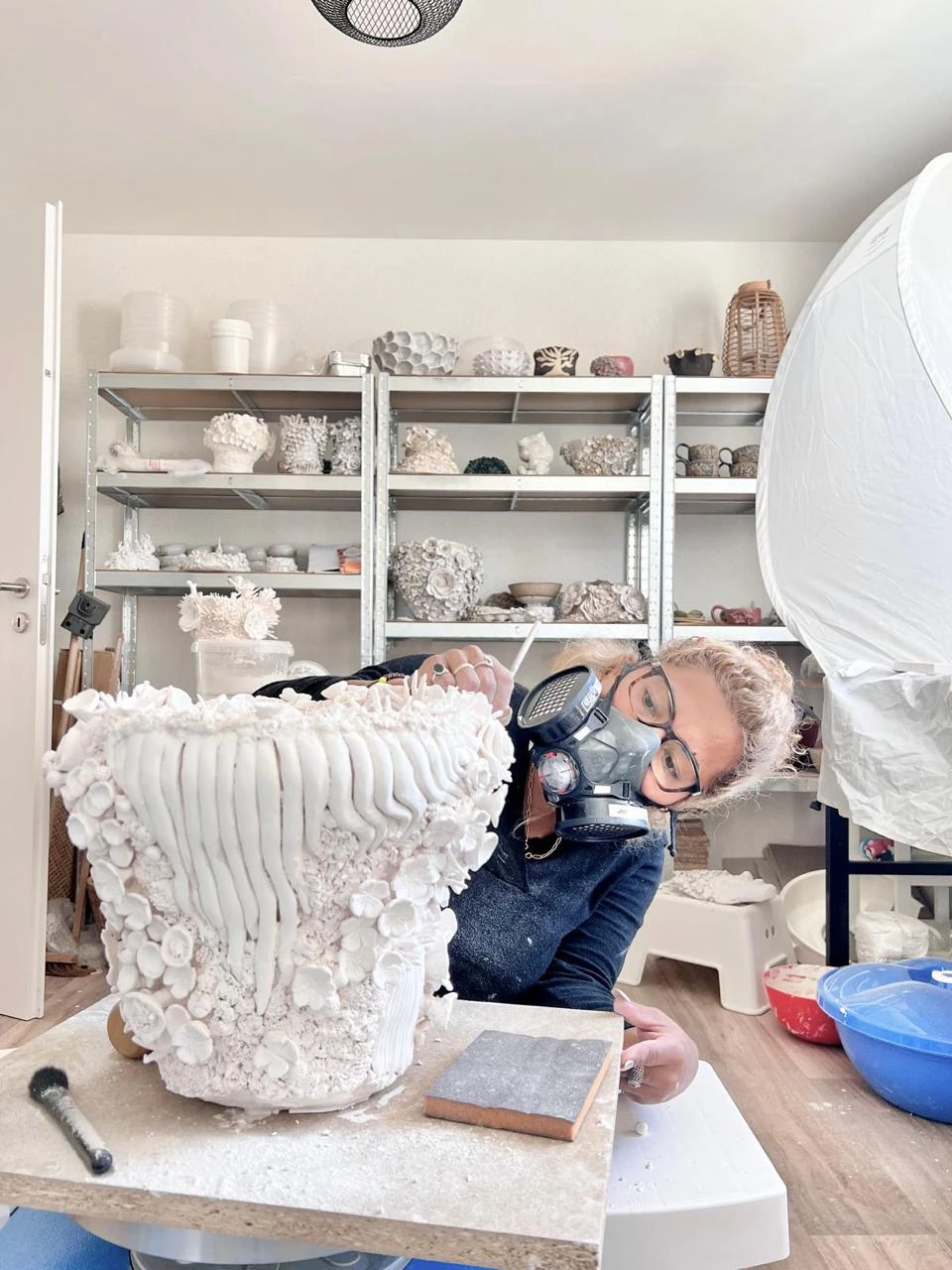 Shristi refining a large floral ceramic vessel at her studio workbench, wearing glasses and a respirator